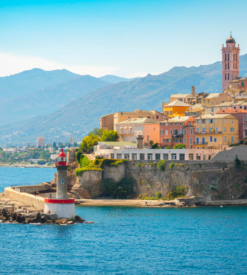 Phare de Bastia sur la côte corse; maisons colorées, mer méditerranéenne et montagnes verdoyantes.