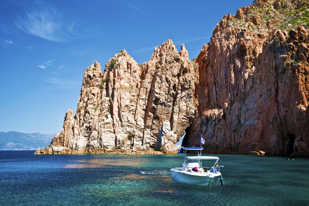 Bateau au large des calanques de Piana en Corse, naviguant sur une mer turquoise sous le ciel ensoleillé. Paysage impressionnant de formations rocheuses, symbole de la nature sauvage corse.