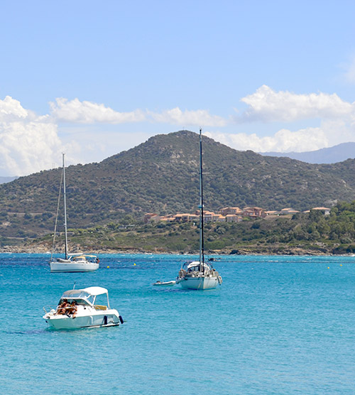 Bateaux naviguant dans la mer turquoise de Corse sous un ciel bleu lumineux, entourés de collines verdoyantes.
