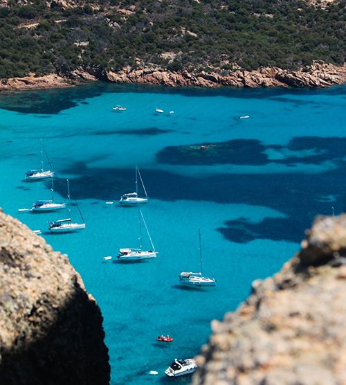 Cette image de bateaux à voile sur une mer bleu éclatante et entourée de côtes corses verdoyantes évoque la beauté naturelle et la sérénité de cette île méditerranéenne ensoleillée.