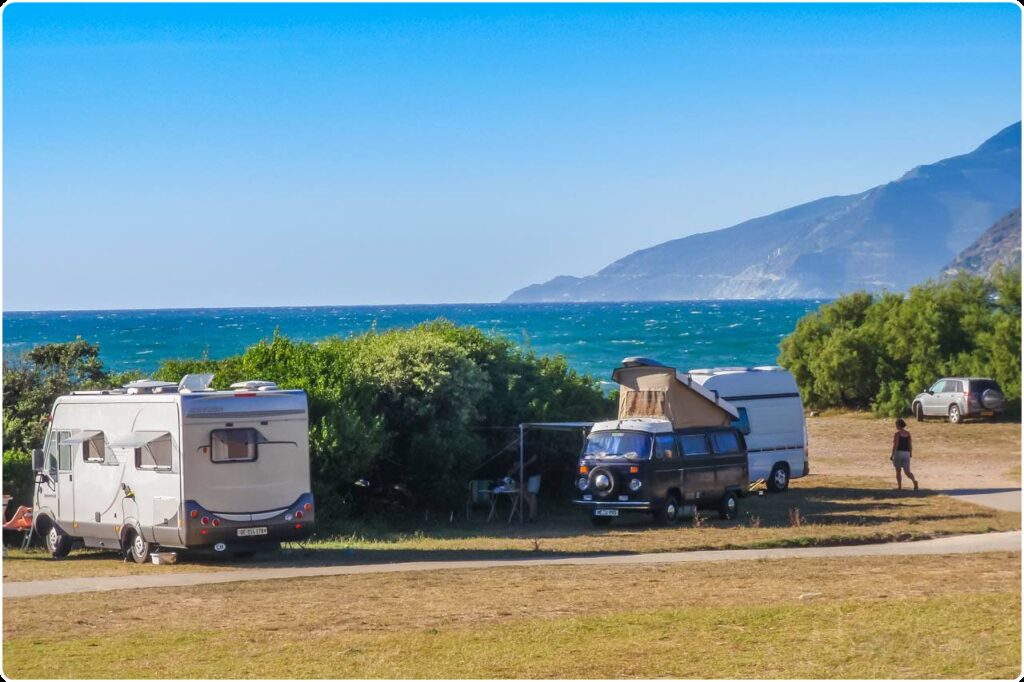 Cette image dépeint des vans en camping face à la mer, sur un magnifique fond de montagnes et de ciel bleu, créant une ambiance de tranquillité et d'évasion en pleine nature.