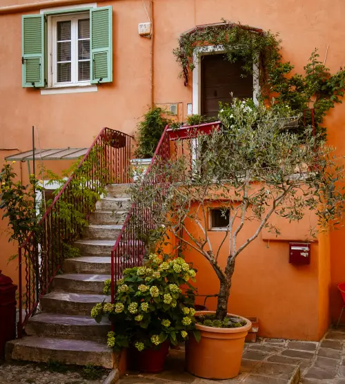 Dieses Bild zeigt eine malerische Treppe vor einem pfirsichfarbenen Haus mit grünen Fensterläden und einem Blumentopf, wodurch eine ruhige, mediterrane Atmosphäre entsteht.