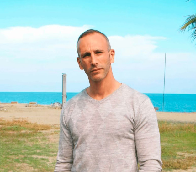 Homme moderne au bord de l'eau sur une plage ensoleillée sous un ciel bleu, incarnant la sérénité et la confiance tranquille.