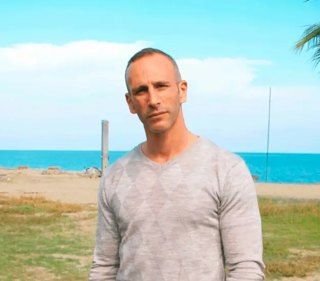 Homme moderne au bord de l'eau sur une plage ensoleillée sous un ciel bleu, incarnant la sérénité et la confiance tranquille.