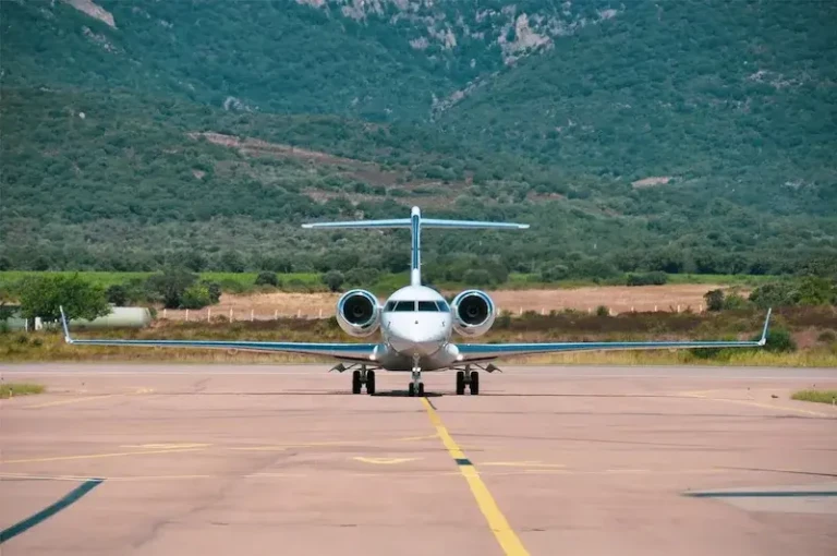 Un avion de type jet privé blanc, vu de face, atterrit ou s'apprête à décoller sur une piste d'aéroport en Corse, avec des montagnes boisées en arrière-plan.