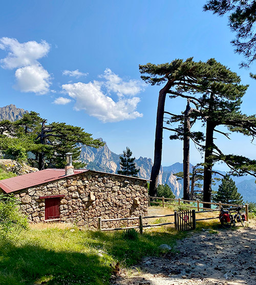 Maison en pierre sur colline, montagnes et ciel bleu en arrière-plan, atmosphère estivale sereine.