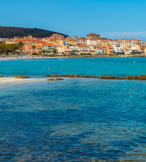 L'image dépeint le village pittoresque de L'Île Rousse avec ses plages dorées baignant dans des eaux cristallines sous un ciel bleu, capturant la sérénité méditerranéenne et l'ambiance estivale.