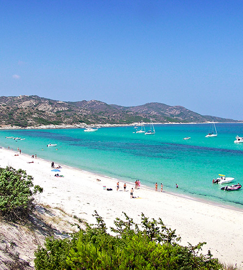 Image d'une plage en Corse avec du sable blanc, mer turquoise et voiliers à l'horizon, capturant une ambiance d'été paisible.