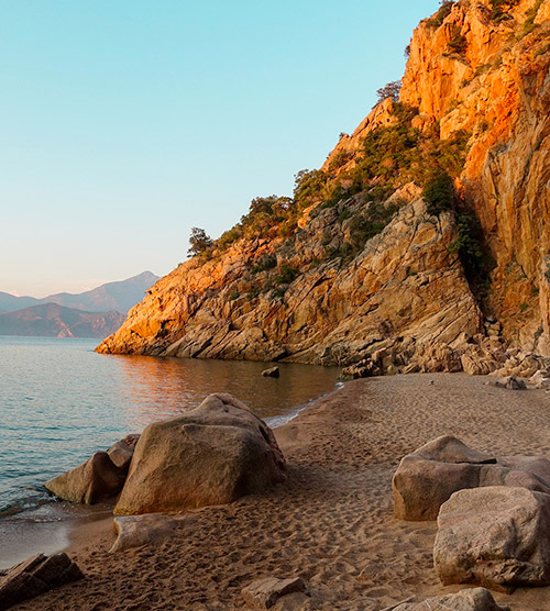 Vue sereine d'une plage au coucher du soleil avec des rochers imposants et des montagnes en arrière-plan. Le sable doré et la lumière crépusculaire créent une atmosphère apaisante, idéale pour se détendre et contempler la nature.