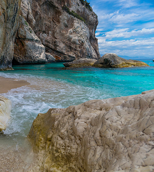 Cette image montre une plage sereine avec des rochers, de l'eau turquoise scintillante, et un ciel bleu, offrant une atmosphère de calme et d'évasion.