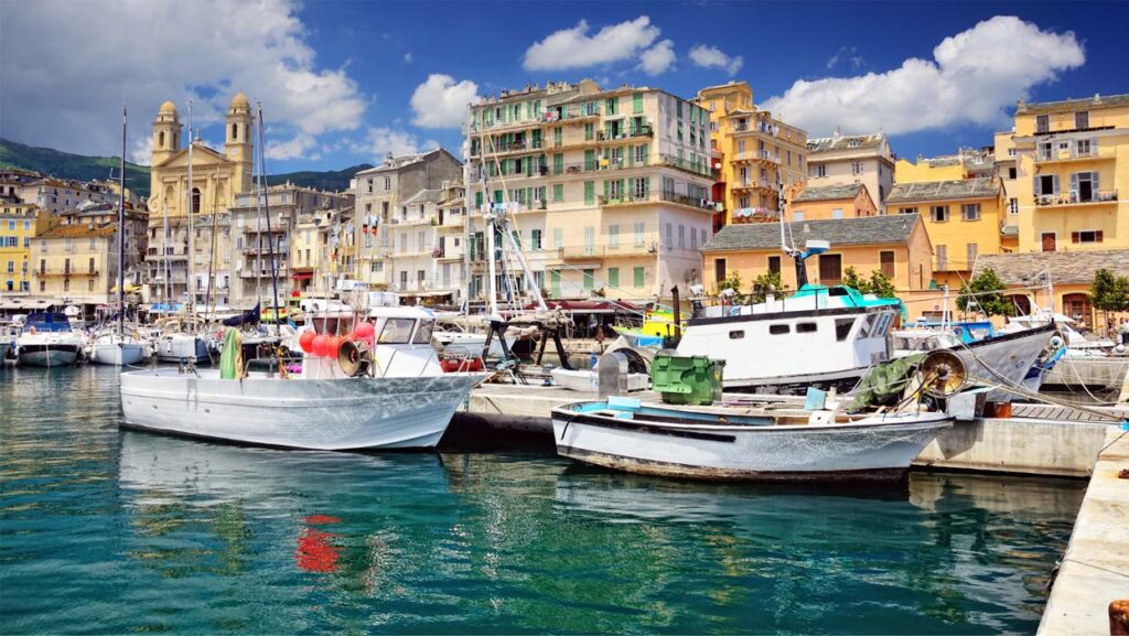 Vieux port ensoleillé avec bateaux de pêche colorés alignés sur les quais, entourés de bâtiments aux façades vibrantes et reflet sur l'eau calme.