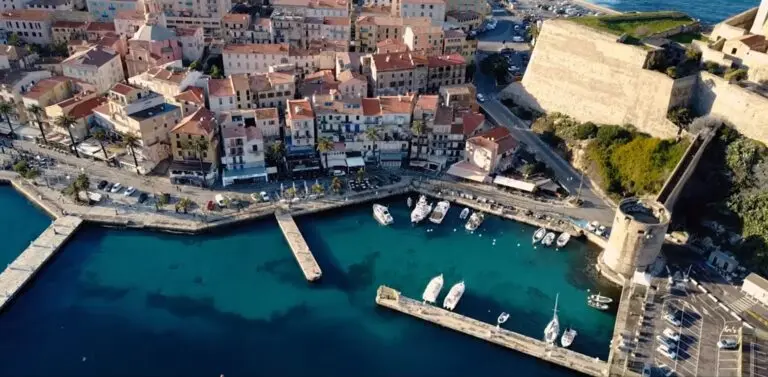 Image aérienne du port de Calvi avec ses voiliers et bâtiments pastel, bordant la Méditerranée, sous le regard d'une tour historique.