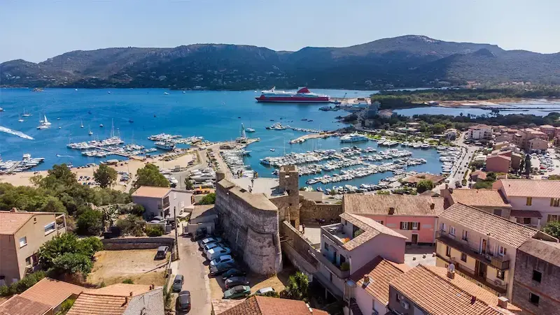 porto-vecchio-vue-aerienne-port-plaisance-commerce-corse Vue aérienne en plongée de la ville de Porto-Vecchio, son port de plaisance rempli de bateaux, un ferry rouge au quai et les montagnes en arrière-plan.