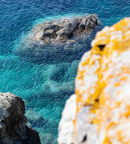 Vue aérienne d'un rocher émergeant d'une mer émeraude, où les nuances de bleu et les reflets lumineux créent une atmosphère apaisante et vivante, soulignant la beauté naturelle de cette scène maritime.