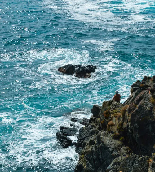 Un homme solitaire se tenant sur des rochers au bord de la mer, symbolisant la solitude et la méditation face à l'immensité de l'océan.