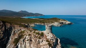 Image panoramique de la Côte Corse avec une tour sur une falaise surplombant la mer, illuminée par le soleil.