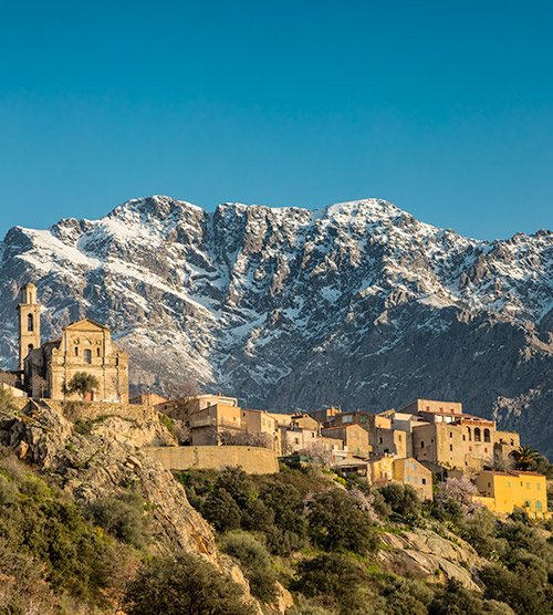 Village corse pittoresque avec montagnes enneigées, ciel bleu, et architecture méditerranéenne.