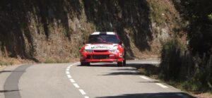 Sur une route montagneuse en Corse, une voiture de rallye rouge exécute un virage serré, illustrant la passion des courses automobiles touchant à la fois à la vitesse et à la beauté des paysages naturels corses.