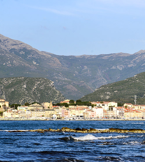 Image d'un paysage côtier en Corse avec un village coloré, montagnes majestueuses et mer cristalline.