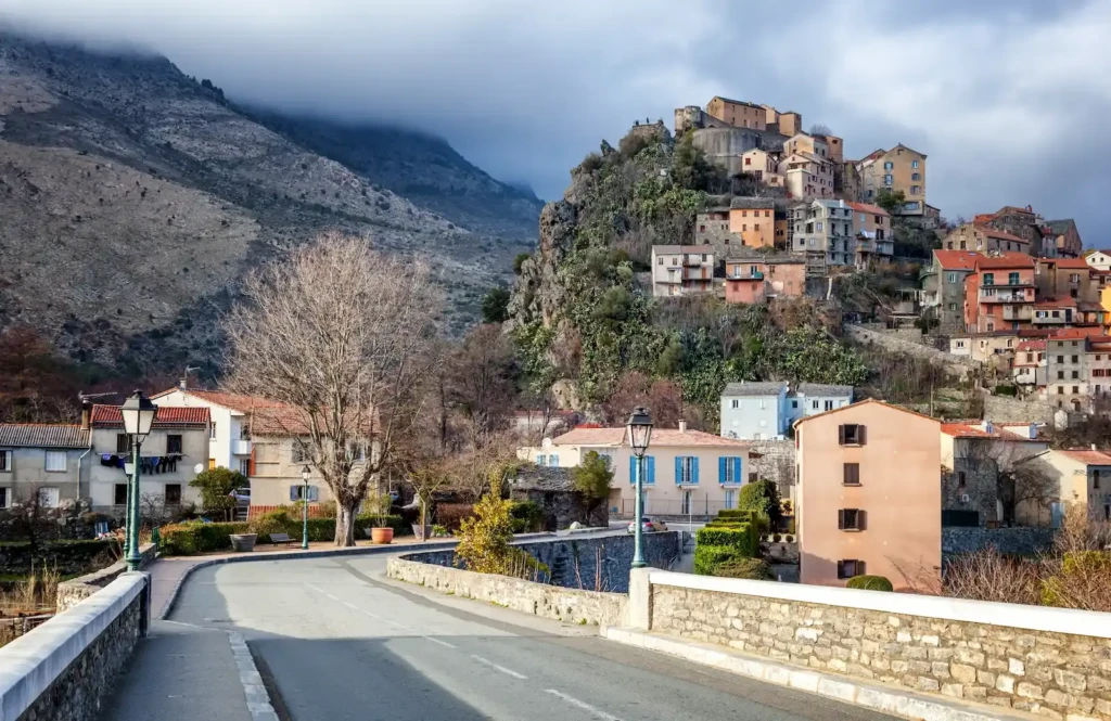 route-et-train-vers-corte Route goudronnée menant vers le village perché de Corte en Corse, avec ses maisons traditionnelles construites sur une colline rocheuse escarpée, surmontée d'une citadelle sous un ciel couvert.