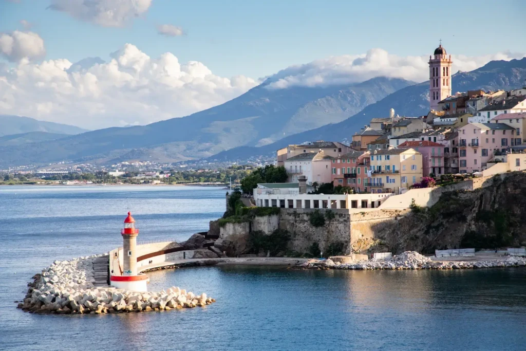 vue-cotiere-sur-la-ville-de-bastia Vue côtière de la ville de Bastia en Corse avec son phare blanc et rouge sur une digue en pierre, ses maisons colorées à flanc de colline et les montagnes en arrière-plan sous un ciel nuageux.