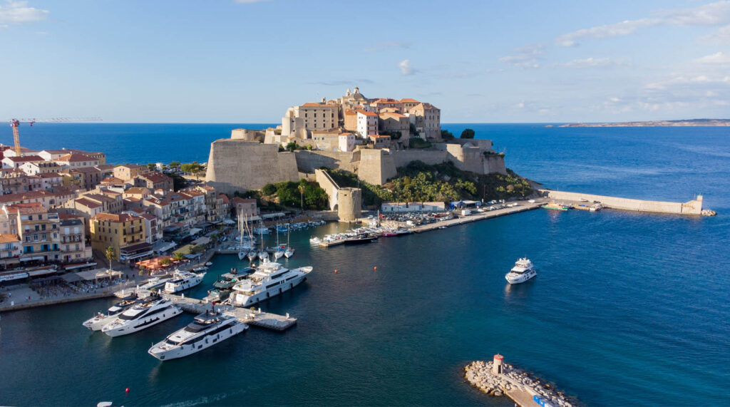 L'image capture la beauté de Calvi avec ses bâtiments colorés et son port historique, évoquant une atmosphère paisible et riche en culture, idéale pour ceux qui souhaitent découvrir la splendeur méditerranéenne et l’histoire en un seul regard.