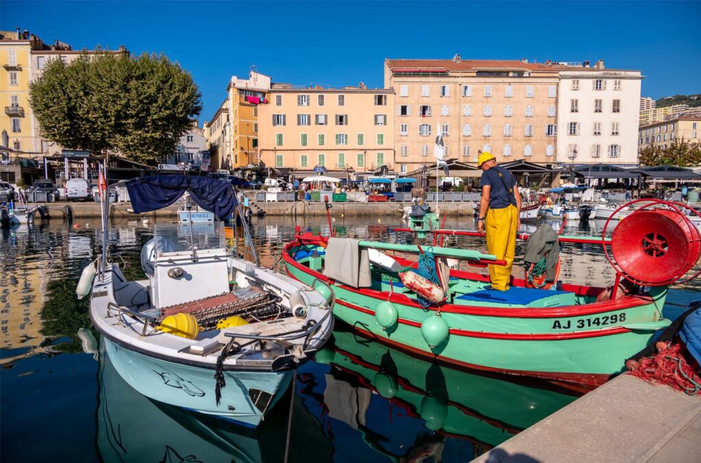 Dans le port d'Ajaccio, un pêcheur sur son bateau coloré sous un ciel azur reflète l'esprit maritime local, entouré d'une ambiance vibrante et sereine.