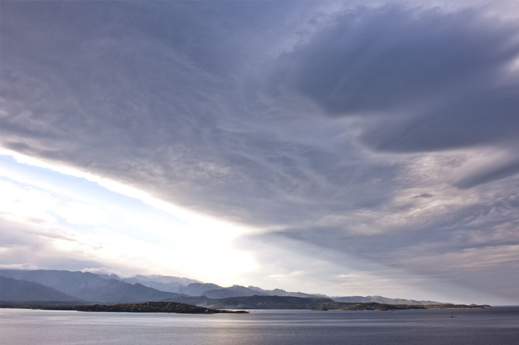 Photographie dramatique d'un ciel nuageux s'étendant sur la mer en Corse, encadrée par des montagnes majestueuses.
