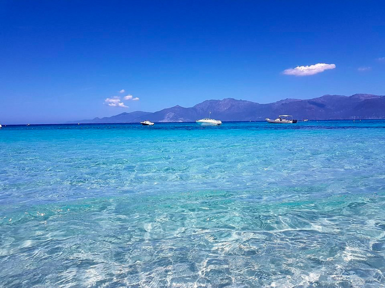 Cette photo montre une plage tropicale avec des eaux cristallines, des bateaux voguant doucement sur une mer bleue, et des montagnes en arrière-plan sous un ciel clair, parfaite pour ceux qui cherchent un cadre naturel éloigné et apaisant.
