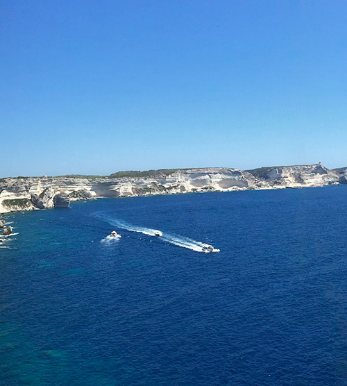Paysage côtier corse avec mer turquoise, bateaux et falaises blanches sous un ciel bleu éclatant, évoquant liberté et aventure.