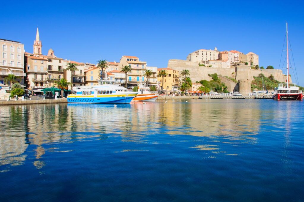 Cette image captivante montre le port de Calvi baigné de lumière, avec ses bateaux colorés flottant sur l'eau turquoise, surplombé par la citadelle, pour une ambiance méditerranéenne idéale.