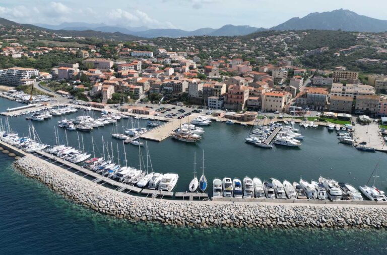 Cette image montre le port de Propriano en Corse sous un jour idyllique. Des yachts étincelants, entourés par des maisons colorées, créent une atmosphère de détente. Les montagnes ondulantes et les reflets solaires sur la mer ajoutent une touche de sérénité méditerranéenne.