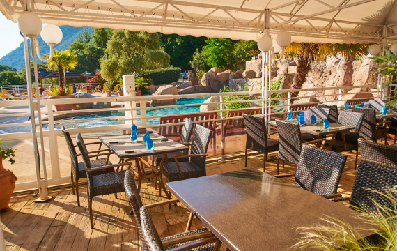 Cette image présente une terrasse de restaurant ensoleillée au bord d'une piscine, idéale pour une expérience culinaire relaxante entourée de verdure.