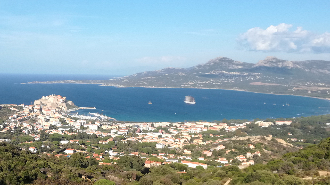 Vue splendide du port corse avec bâtiments pittoresques et mer méditerranéenne sous un ciel bleu clair, idéal pour capturer l'essence sereine et invitante de l'île.