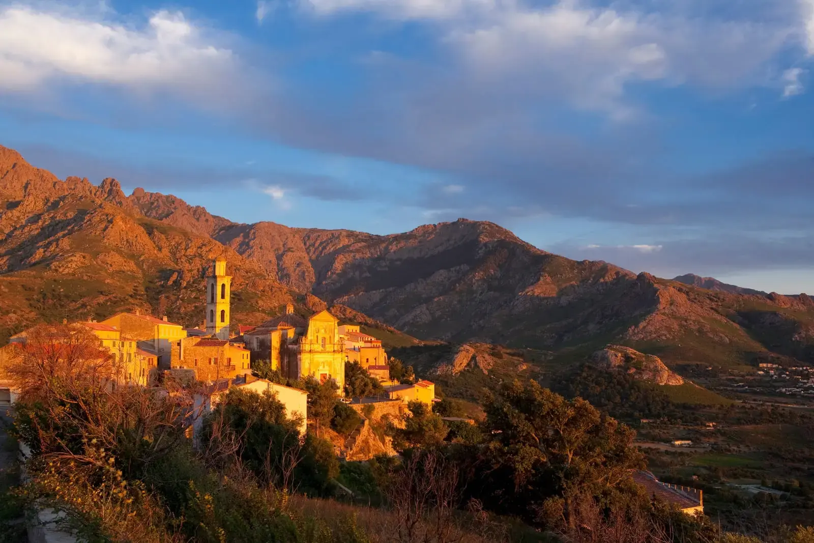 Le village perché de Speloncato en Balagne, baigné par la lumière dorée du soleil couchant. On distingue l'église paroissiale San Michele et son clocher au cœur des maisons en pierre, avec des montagnes arides en arrière-plan.