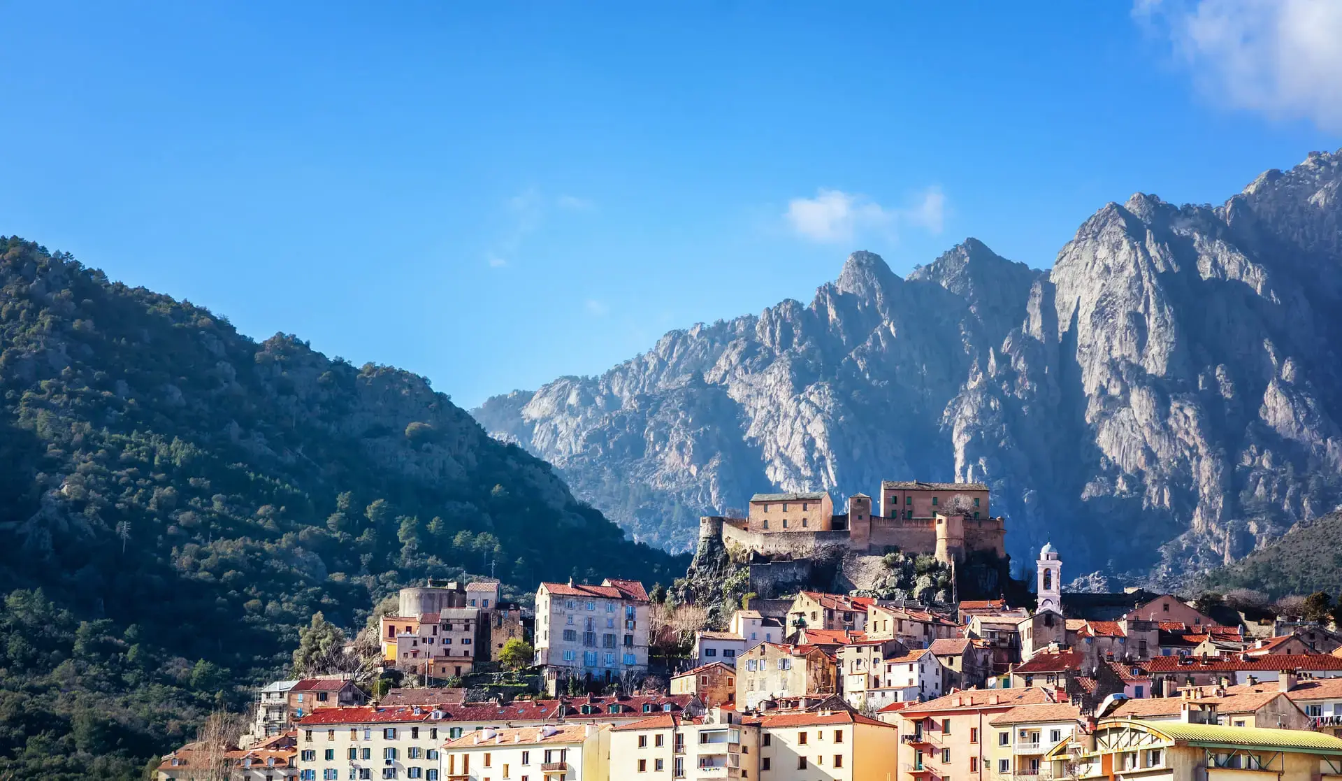 Vue sur le village de Corte avec ses maisons aux toits de tuiles oranges, dominé par sa citadelle historique perchée sur un éperon rocheux, avec en arrière-plan les sommets granitiques imposants de la vallée de la Restonica sous un ciel bleu.