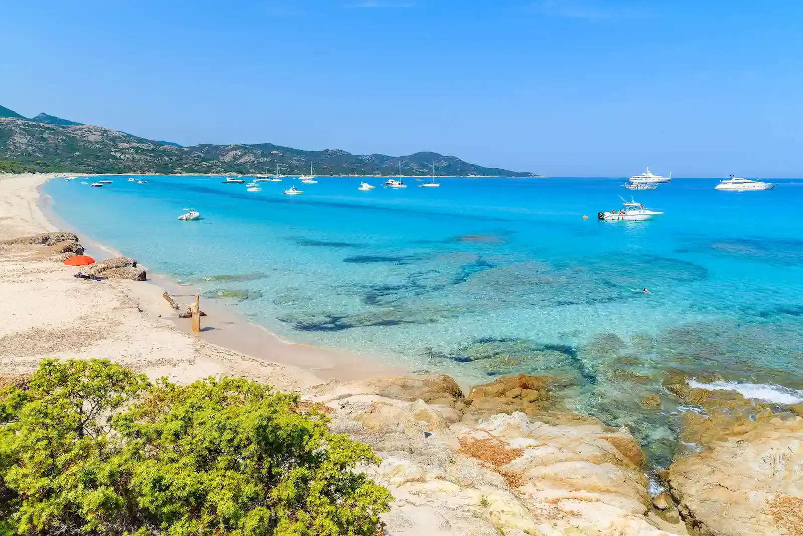 Eaux turquoise et sable fin de la plage du Lotu (Loto) dans le désert des Agriate. Des bateaux de plaisance mouillent dans la baie bordée de collines sauvages sous un ciel azur.