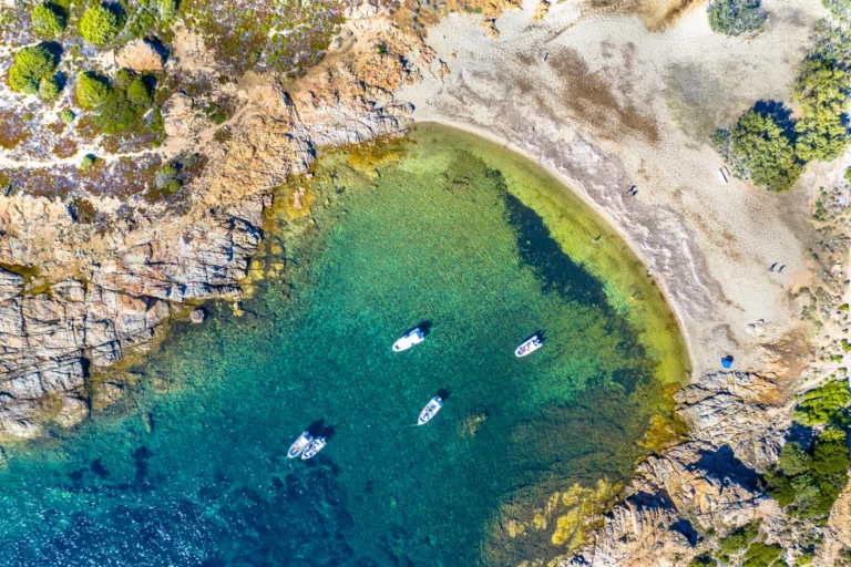 Vue aérienne d’une crique corse aux eaux turquoise avec plusieurs bateaux amarrés près du rivage sableux.