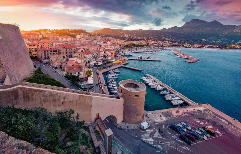 Quand partir en Corse en 2026 Vue aérienne du port de Calvi en Corse au coucher du soleil, avec la citadelle surplombant la mer.