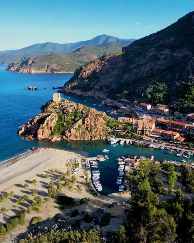 Port-Ota-Porto-Corse-Vacances-Balade-Bateau-Pass-Partout Vue aérienne du golfe de Porto avec sa tour génoise perchée sur un rocher, la marina avec ses bateaux, la plage de galets et les montagnes environnantes.