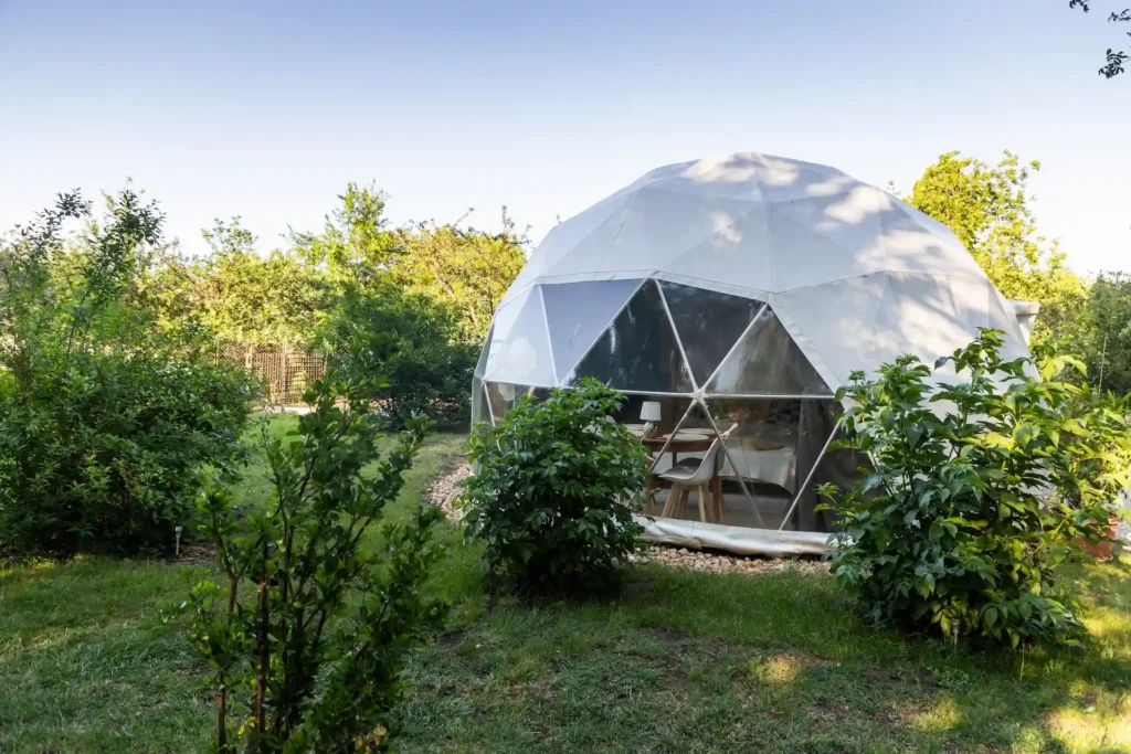 hébergement-insolite-corse-bulle-3-étoiles Dôme géodésique blanc et transparent installé sur un terrain engazonné bordé d'arbustes et de verdure.