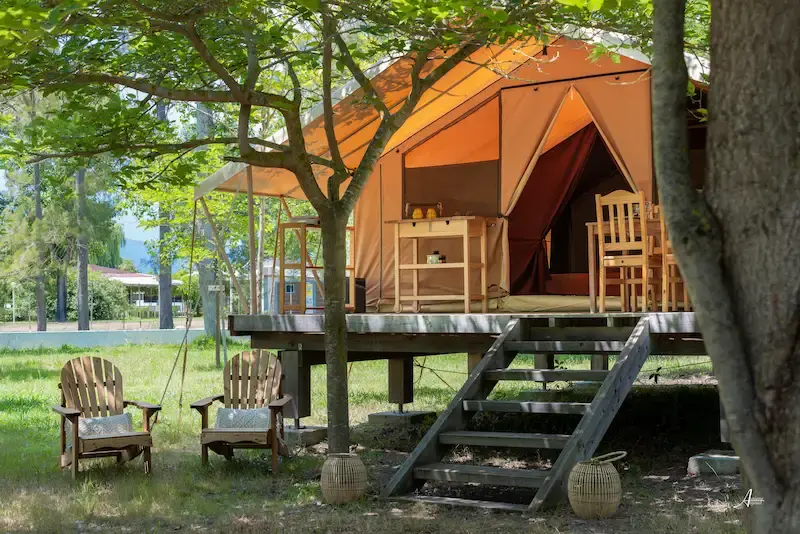 logement-insolite-au-camping-u-casone Tente lodge safari en toile orange installée sur une terrasse surélevée en bois avec salon de jardin sous les arbres.