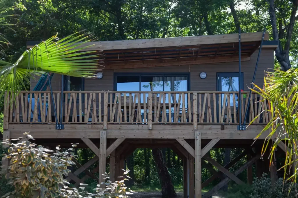 pilotis-hebergement-insolite-en-corse-merendella Cabane contemporaine en bois sur pilotis avec grande baie vitrée et terrasse surélevée entourée de palmiers et d'arbres.